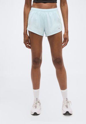 Γυναίκα με ανοιχτό μπλε αθλητικά σορτς, λευκές κάλτσες και λευκά αθλητικά παπούτσια, στέκεται με τα πόδια λίγο ανοιχτά πάνω σε λευκό φόντο.