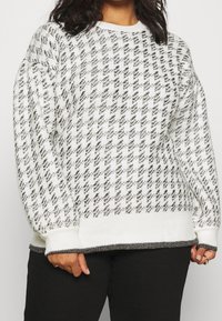 Femme portant un pull en tricot à motif pied-de-poule noir et blanc avec des poignets et un ourlet côtelés, associé à un pantalon noir.