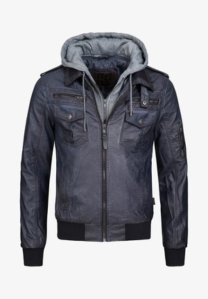 Mann trägt schwarze Lederjacke über grauem Hoodie, weißem Rollkragenpullover, hellgrauen Jogginghosen und schwarzen Sneakers und steht vor einem einfarbigen Hintergrund.