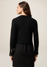 Zwarte cropped jack van gevarieerd materiaal, met accentuering op de mouwen voorzien van gouden knopen en een gegolfde onderrand. Zichtbaar ontwerp aan de achterkant.