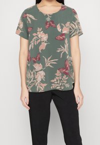 Mujer vistiendo una blusa verde de manga corta con patrones de flores y mariposas rosas, combinada con pantalones negros, de pie contra un fondo blanco.