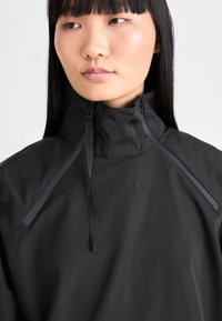Schwarze Zip-Jacke mit hohem Kragen, ausgestattet mit matter Oberfläche, verstellbaren Kordeln und subtiler Markenkennzeichnung auf der Brust.