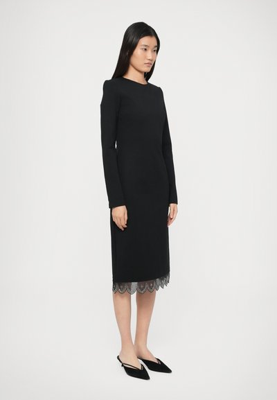Fekete hosszú ujjú ruha, testhezálló sziluettel, hullámos szegélyű csipkecsipkével. Az anyag sima és formázott hatású.