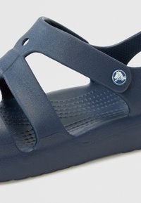 Námorně modré gumové sandály s křížovým designem pásků, texturovanou stélkou a kruhovým detailem loga na pásku. Hladký povrch a nízká hmotnost.