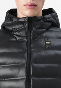 Zwart pufferjack met capuchon, met een gewatteerde textuur, vooraanrits en een logopatch op de linkerborst. Glad, synthetisch materiaal.