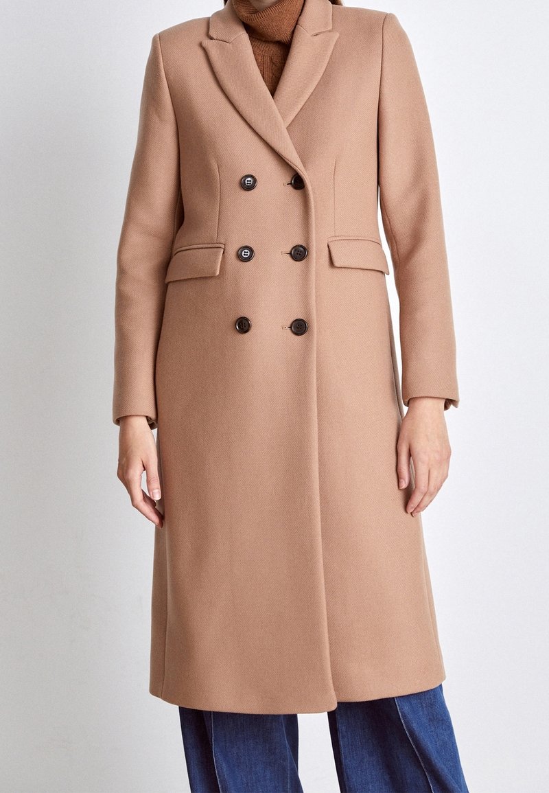 Femme portant un long manteau croisé couleur chameau par-dessus un pull à col roulé marron et un jean bleu sur un fond clair uni.