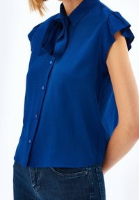 Blauwe blouse met korte mouwen, een kraag en een strikdetail. Gladde stof, knoopsluiting aan de voorkant en subtiele gerimpelde mouwen.