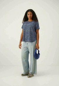 Mulher com cabelo longo e encaracolado, vestindo uma blusa azul com flores, jeans largos de perna clara, sapatilhas, segurando uma mala azul contra um fundo simples.