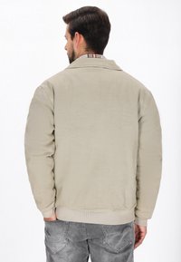 Lys beige kordfløjlsjakke med krave, rillede manchetter og talje. Bagsiden har en glat tekstur med subtile linjer og sømme.