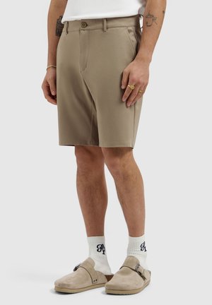 Person trägt beige kniehohe Shorts, weiße Socken mit schwarzer Schrift, beige Wildleder-Slipper und ein weißes T-Shirt.