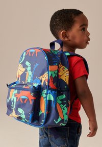 Mladý chlapec v červenej košeli a džínsoch s farebným batohom s motívom dinosaurov na svetložltom pozadí.