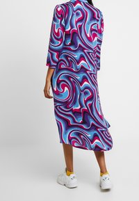 Kolorowa sukienka z długim rękawem z wirującym wzorem w niebieskim, różowym i purpurowym kolorze. Połączona z białymi sneakersami i żółtymi skarpetkami.