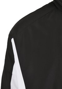 Gros plan sur l'épaule d'une veste noire avec un détail de bande verticale blanche le long de la manche, montrant la texture du tissu et les coutures.