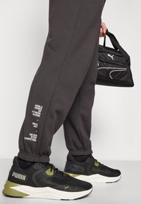 Schwarze Netz-Sportschuhe mit olivfarbenen Akzenten. Graue Jogginghose mit weißen Schriftzügen. Kompakte schwarze Tragetasche mit Logo.