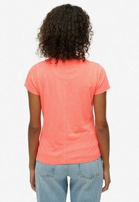 Koraalkleurig T-shirt met korte mouwen en een ronde halslijn, gladde textuur en subtiele middennaad, gecombineerd met lichtblauwe jeans.