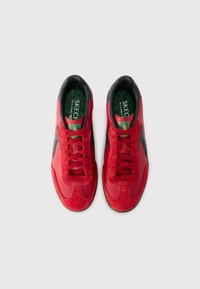 Baskets rouges présentant un mélange de textures lisses et en daim, des accents noirs, un design à lacets, et des semelles intérieures vertes avec un détail logo.