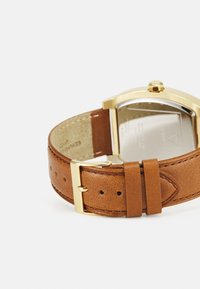 Pulseira de relógio em couro castanho com fivela em metal dourado, apresentando uma textura suave e detalhes de costura visíveis. Design minimalista com caixa retangular.