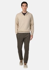 Haut à col zippé beige avec une coupe décontractée, associé à un pantalon plissé gris foncé et des baskets beiges avec des accents marron.