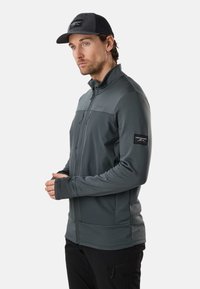 Homme portant une veste zippée gris foncé et une casquette noire, debout les mains jointes, regardant légèrement sur le côté sur un fond uni.