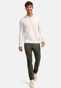 Camisa polo de manga larga en un beige claro, con cuello y detalles de botones, combinada con pantalones verdes oscuros y zapatillas beige, con un corte relajado.