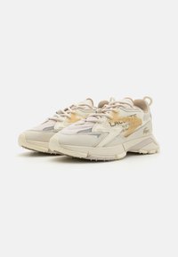 Baskets Lacoste en crème avec des accents beige clair, présentant un dessus texturé, des matériaux contrastants et une semelle en caoutchouc cramponnée.