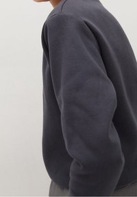 Tmavě šedá mikina vyrobená z měkké tkaniny, s kulatým výstřihem a dlouhými rukávy, zobrazená z boku s mírným texturováním.