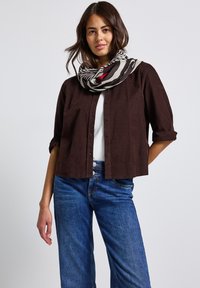 Donna che indossa una camicia button-down marrone scuro con maniche arrotolate, una maglietta bianca, jeans blu e una sciarpa a righe bianco e nero con dettagli rossi.