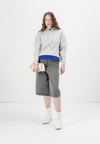 Szürke kapucnis pulóver, amely egy kék felsőre van rétegezve, szürke farmerhotpants és fehér bokacsizma, miközben egy kis krém színű kézitáskát tart.