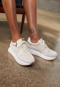 Chaussures de sport beige avec panneaux en maille et accents métalliques, dotées d'une semelle blanche épaisse et de lacets, posées sur un sol texturé.