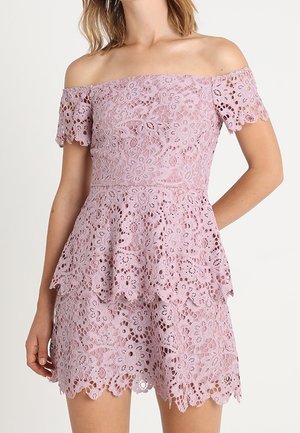 Femme portant une robe en dentelle lilas clair épaules dénudées avec motifs floraux et bords festonnés, avec une jupe à étages et des manches courtes.