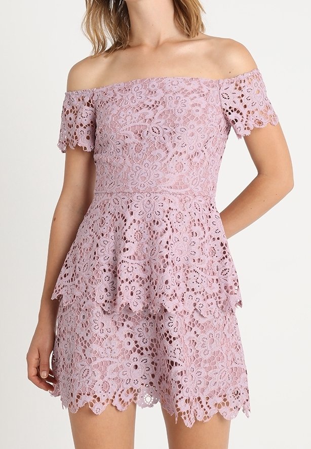 Femme portant une robe en dentelle lilas clair épaules dénudées avec motifs floraux et bords festonnés, avec une jupe à étages et des manches courtes.