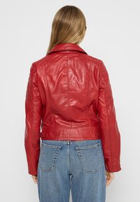 Chaqueta de cuero roja con un diseño ajustado, que presenta un collar puntiagudo y acentos acolchados en los hombros. Combinada con jeans azules de tiro alto.