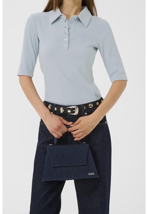 Femme portant une chemise bleu clair boutonnée et un jean foncé avec ceinture cloutée, tenant un petit sac à main bleu foncé devant un fond blanc.