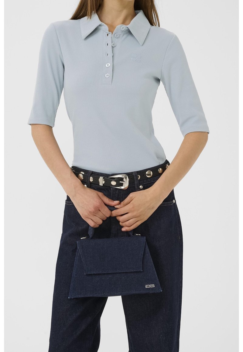 Femme portant une chemise bleu clair boutonnée et un jean foncé avec ceinture cloutée, tenant un petit sac à main bleu foncé devant un fond blanc.