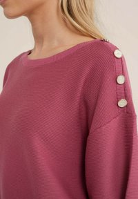 Stickad tröja i en rik rosa färg med rundad halsringning och knappar längs axeln. Texturen är ribbad och något löst sittande.