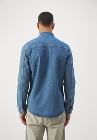 Denim košulja u srednje plavoj boji s dugim rukavima, klasičnom kragnom i zaobljenim rubom. Ima jedan dugme na manžetama i detalje šivanja.
