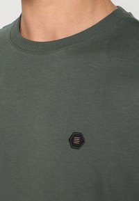 Olijfgroene katoenen trui met een ronde hals. Voorzien van een zwart zeshoekig embleem met drie horizontale gouden strepen. Gladde textuur.