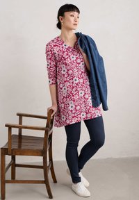 Florale Tunika-Shirt in Burgunderrot mit weißen und schwarzen Blumen. Kombiniert mit dunklen Jeans und weißen Sneakers, mit einer Jeansjacke über dem Arm.