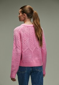 Roze gebreide trui met een textuurtje patroon, met een ronde halslijn en geribbelde manchetten. Gecombineerd met een blauwe jeans, die iets in de taille is gestoken.