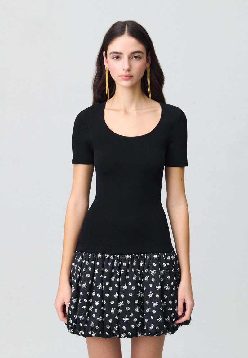 Haut noir ajusté à manches courtes et décolleté en cœur, associé à une jupe noire évasée ornée de motifs floraux blancs.