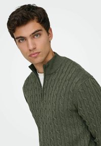 Pull en maille torsadée verte avec un col montant et une fermeture éclair mi-haute, présentant un motif texturé et une coupe ajustée.