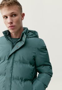 Zelená zateplená bunda s kapucňou, so zipsovým zapínaním a cvokmi. Materiál má hladkú textúru a je prešívaný pre dodatočné zateplenie.