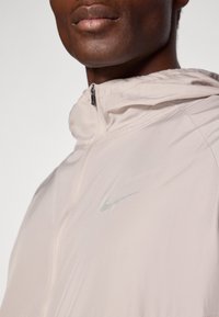 Lys beige jakke med lynlås foran, der har et enkelt design og et lille metallisk Nike-logo på venstre side. Glat tekstur.