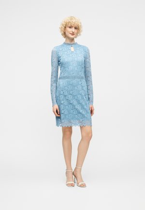 Kobieta z krótkimi kręconymi blond włosami, ubrana w jasnoniebieską koronkową sukienkę z długim rękawem i beżowe sandały na wysokim obcasie, stojąca na białym tle.