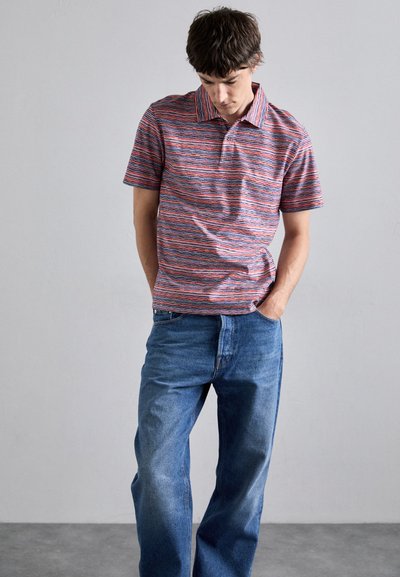 Ριγέ πόλο μπλούζα σε κόκκινο, μπλε και λευκό, με κοντά μανίκια και ριγωτό γιακά. Φοριέται με μπλε τζιν παντελόνι, σε χαλαρή εφαρμογή, με ορατές λεπτομέρειες ραφής.