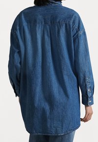 Denimhemd in Mittelblau mit langen Ärmeln und Knopfmanschetten. Verfügt über eine Rückenfalte und einen abgerundeten Saum für eine lässige Passform.