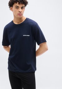 Νεαρός άνδρας με σγουρά μαλλιά, που φοράει μπλε σκούρο μπλουζάκι Jack & Jones και μαύρο παντελόνι, στέκεται μπροστά σε απλό φωτεινό φόντο.