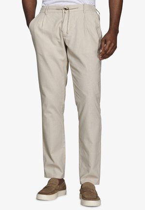 Mężczyzna ubrany w beżowe spodnie chino z wiązaniem w pasie i brązowe wsuwane buty, stojący z jedną ręką w kieszeni.