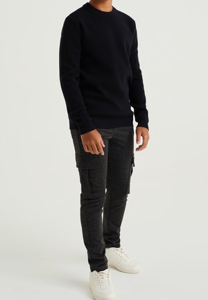 Tummansininen neulepaita, jossa on joustavat hihansuut ja helma, yhdistetty mustiin ruudullisiin cargo-housuihin ja valkoisiin lenkkareihin.