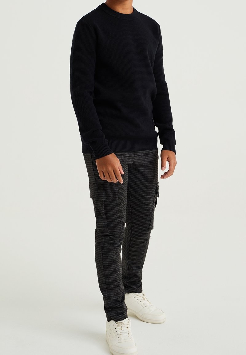 Tummansininen neulepaita, jossa on joustavat hihansuut ja helma, yhdistetty mustiin ruudullisiin cargo-housuihin ja valkoisiin lenkkareihin.